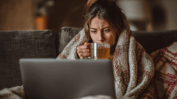 Travailleuse indépendante malade en télétravail à domicile, emmitouflée dans une couverture, tasse de tisane à la main, travaillant malgré la fièvre.